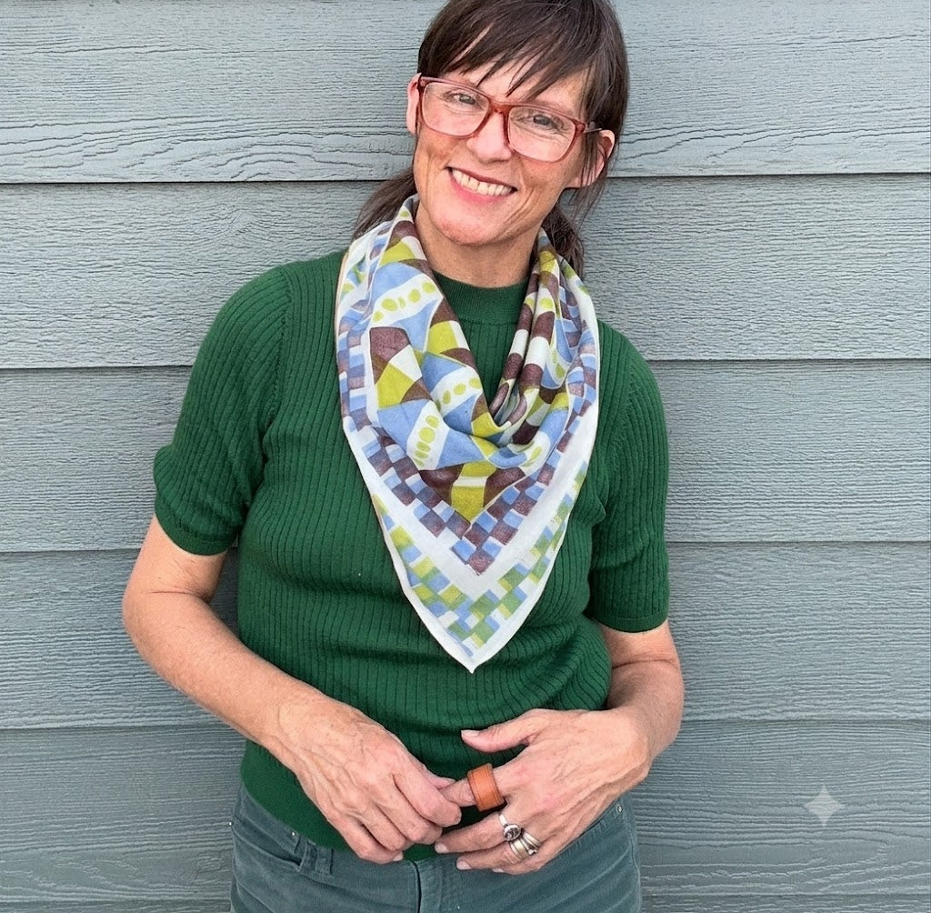 A woman wearing a green top stands against a gray wood-paneled wall, smiling and wearing a geometrically patterned scarf in white, blue, yellow, and brown.