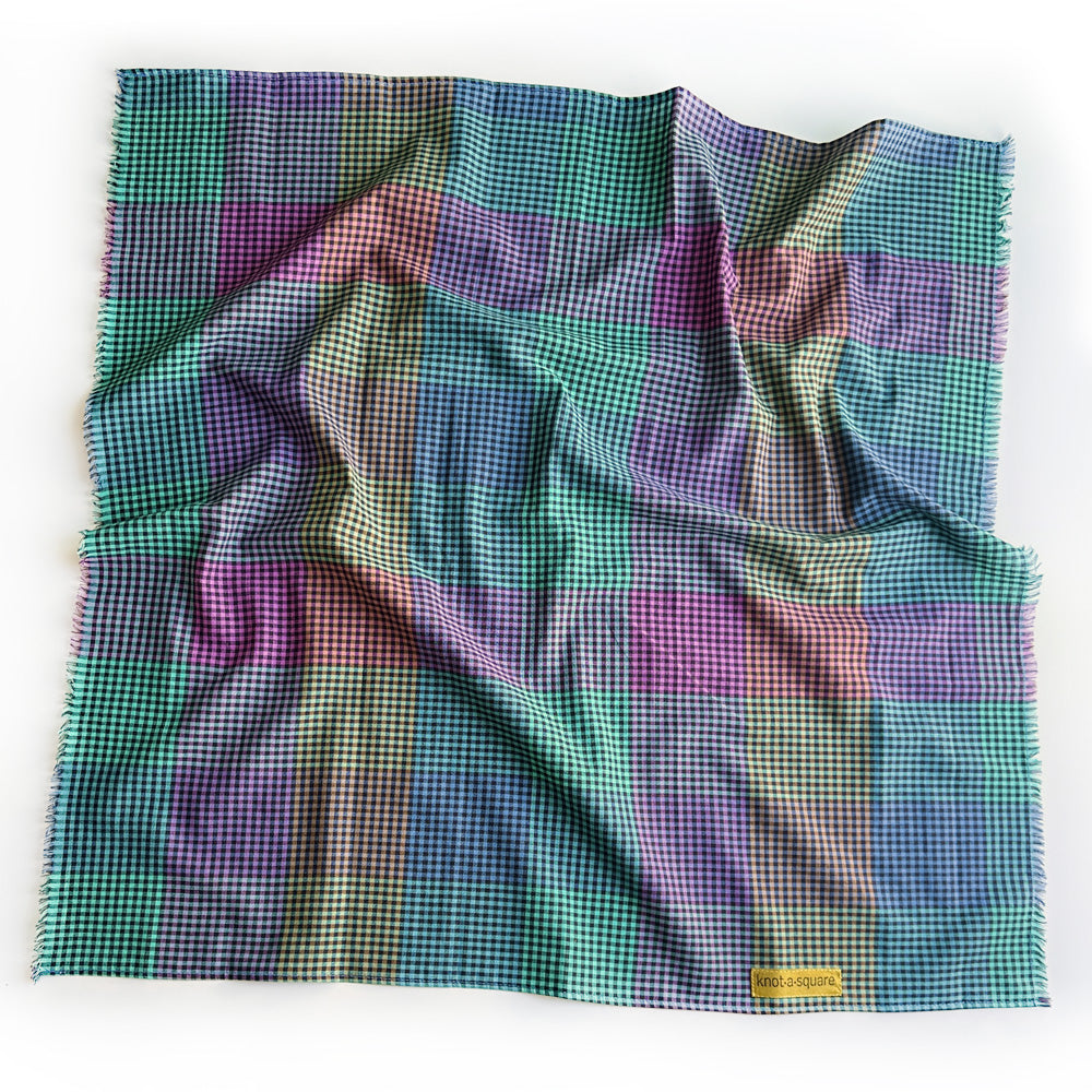 multi color gingham square scarf  with fringed edges on a white background.  The gingham check pattern of teal, purple, green, and blue, featuring a small white brand tag that reads 'KNOT A SQUARE'.