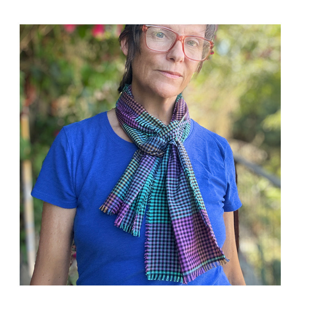 A woman wearing a multi color gingham scarf, in a vibrant array of cool colors.   She is outdoors, surrounded by greenery.