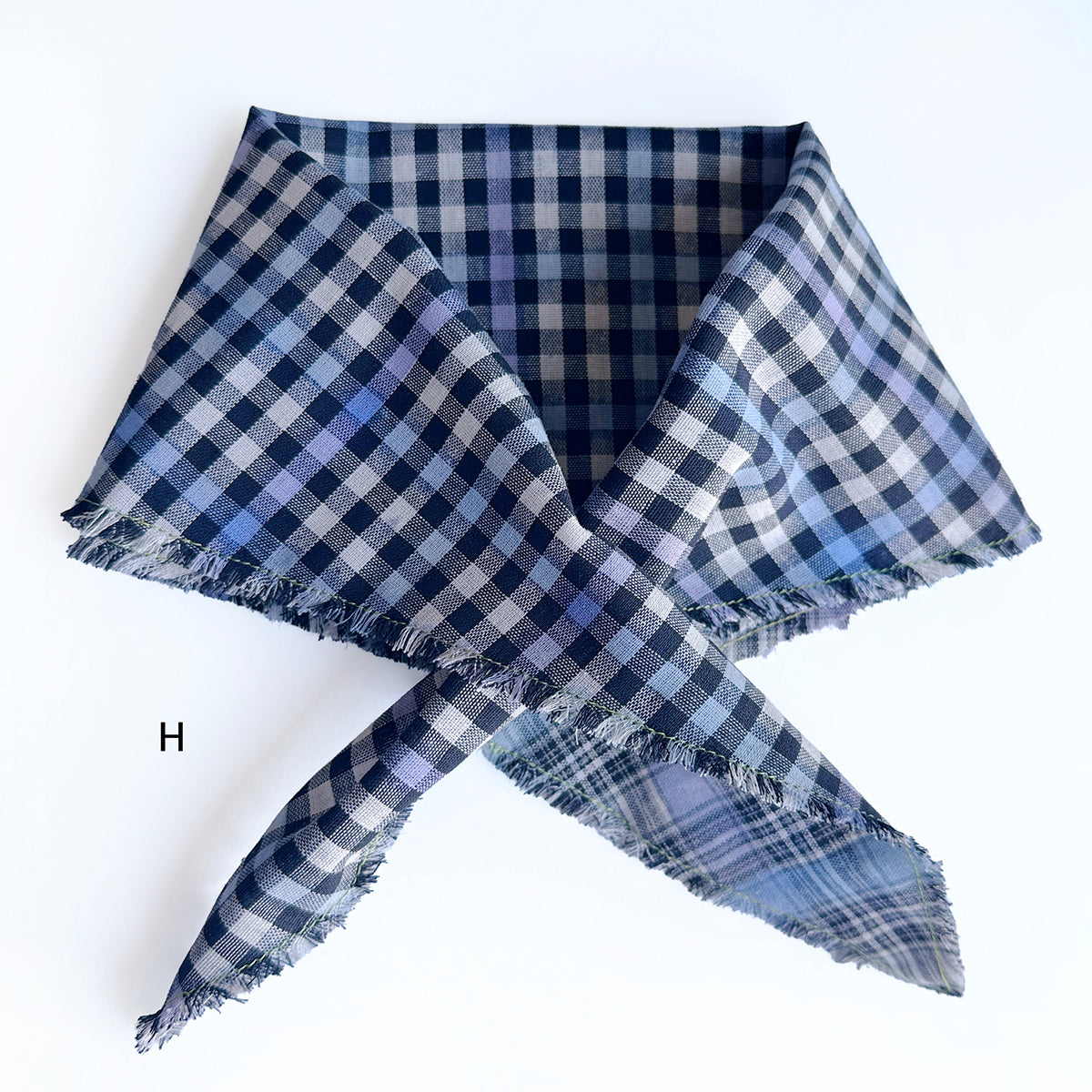 A diamond shaped plaid scarf with frayed edges, folded and tied in a knot on a plain white background. The pattern features navy, blue, and lavender checks, with a small black letter 'H' printed on the left side.