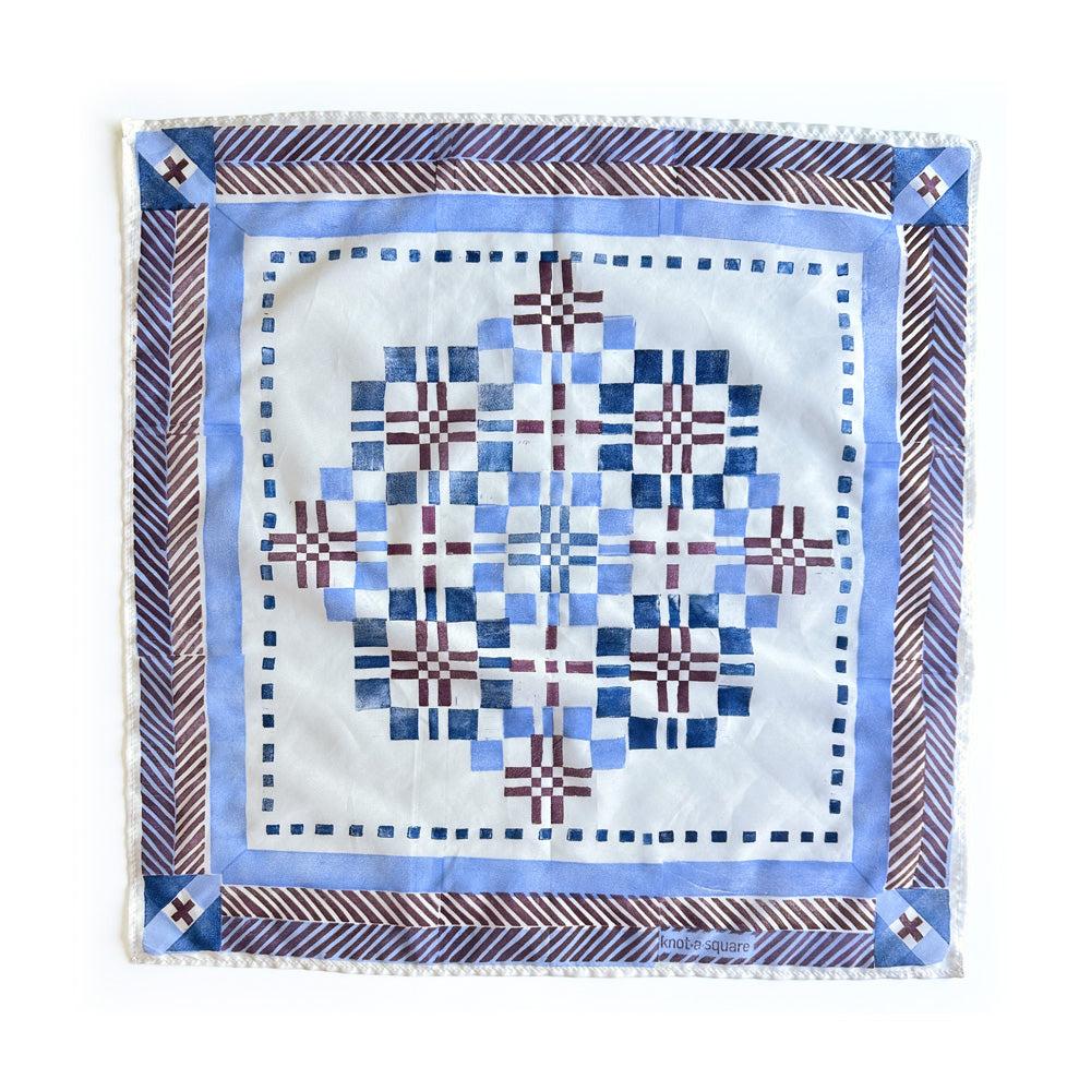 A square cotton silk scarf featuring a geometric patchwork design in shades of blue, white, and garnet, finished with a patterned border.