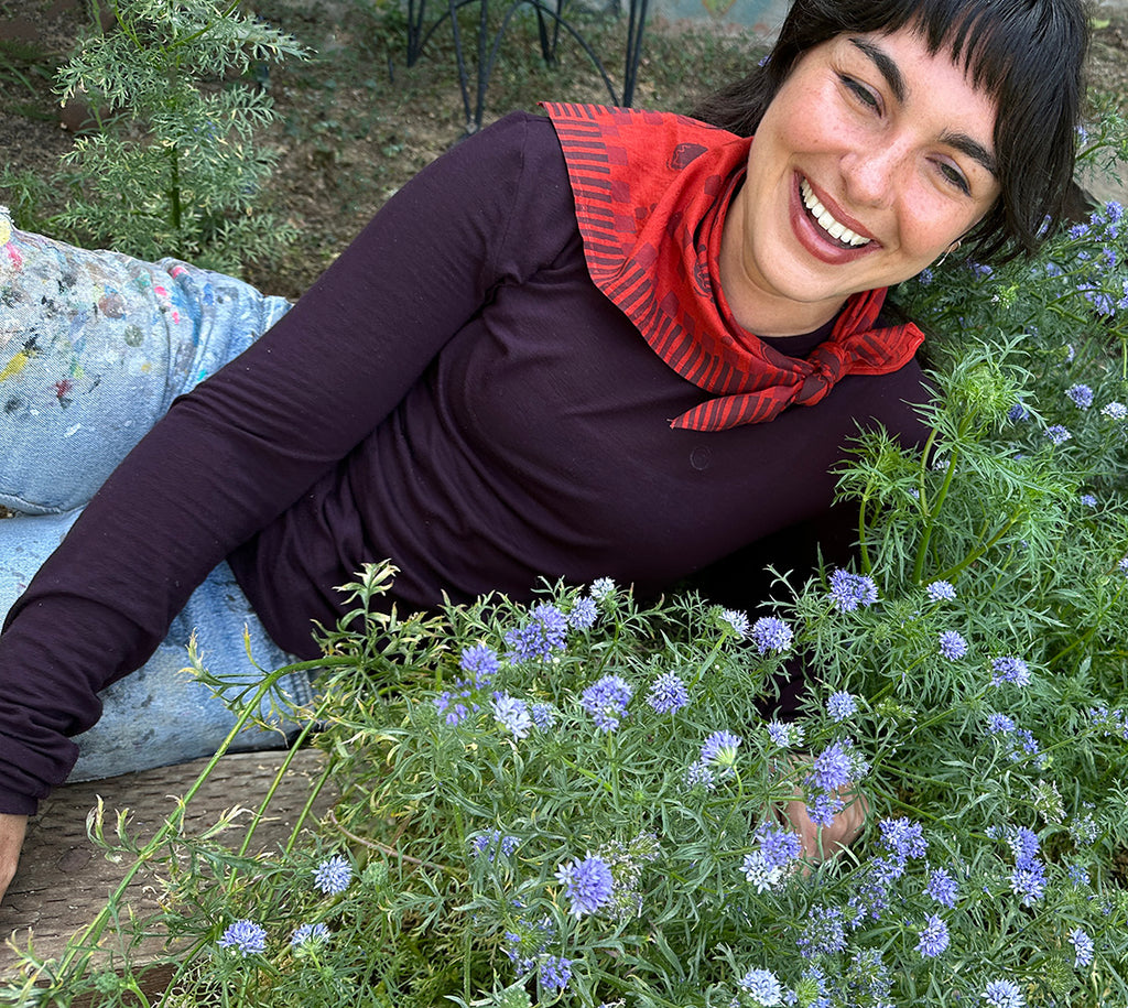 A smiling woman lying among flowers, wearing a hand painted persimmon-red bandana.