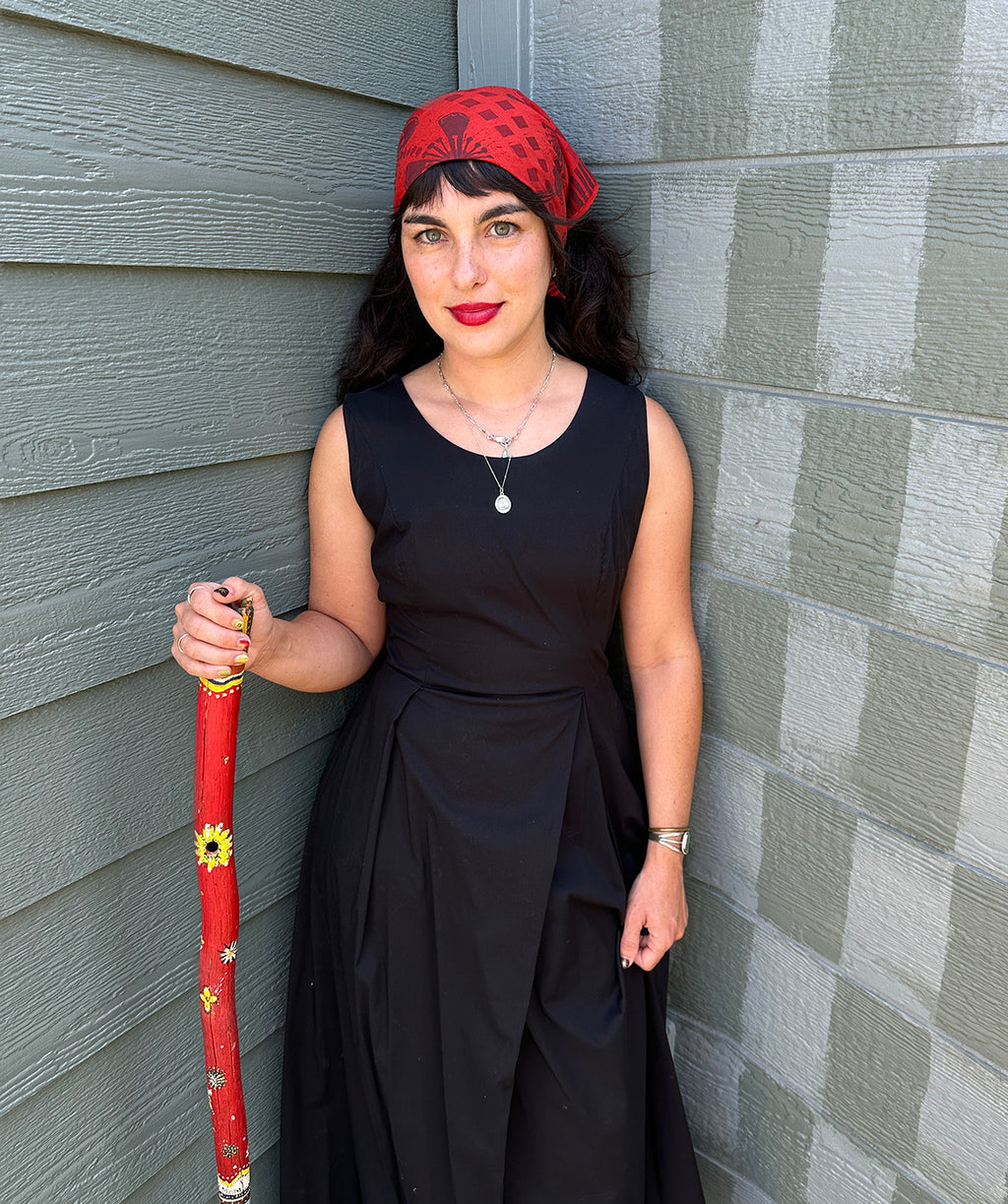 Woman in black dress with red headscarf and walking stick against a gray wall