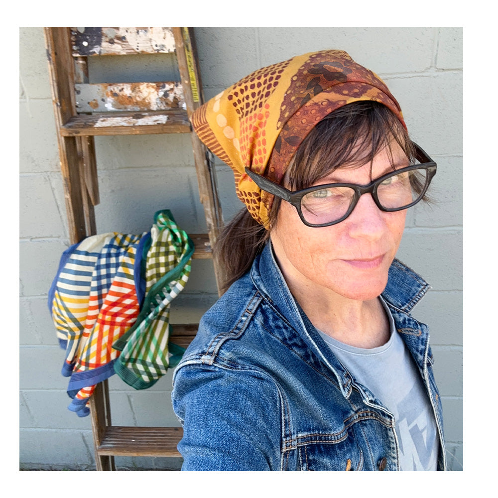 Person wearing a colorful headscarf with a wooden ladder and patterned scarves in the background.
